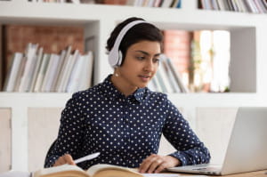 Attractive indian ethnicity girl do assignment preparing for university test or exams seated at desk in public library wearing headphones listen audio task, e-learning process educational app concept